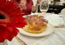 A round-braided challah loaf, on a table surrounded by wine glasses, cutlery, and a red flower.