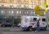 A young girl wearing a helmet and an older person holding a scooter talk to an ice cream truck driver on a city street in front of a large scaffolded building.