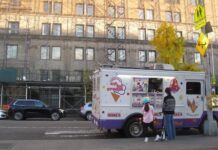 A young girl wearing a helmet and an older person holding a scooter talk to an ice cream truck driver on a city street in front of a large scaffolded building.