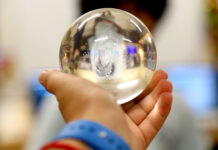 A hand holding a clear glass sphere, with a blurred background and a person’s face partially visible through the sphere.