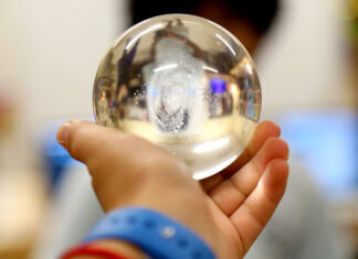 A hand holding a clear glass sphere, with a blurred background and a person’s face partially visible through the sphere.
