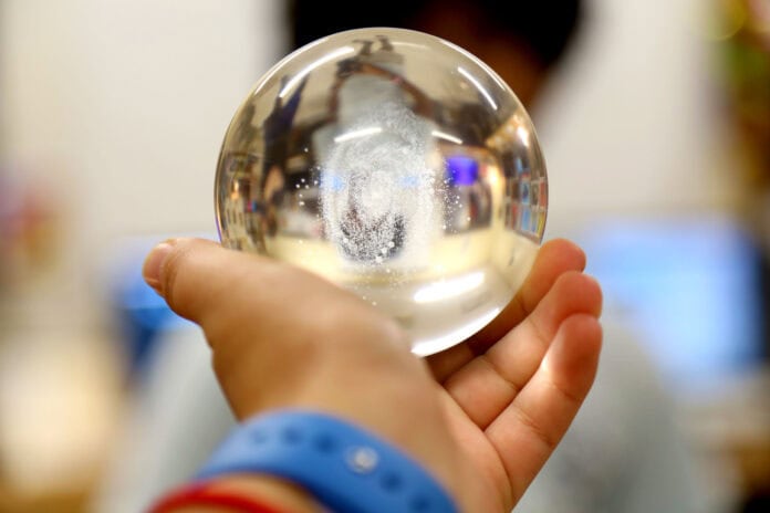 A hand holding a clear glass sphere, with a blurred background and a person’s face partially visible through the sphere.