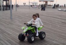 Two young girls ride a green four-wheeler over a boardwalk