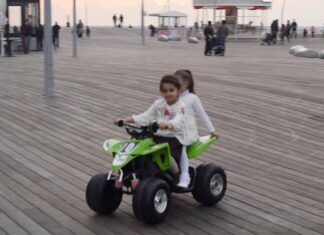 Two young girls ride a green four-wheeler over a boardwalk