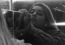 A young woman looks at herself in a scratched mirror, her chin angled up and her expression bored or perhaps sorrowful. The photo is in black and white.