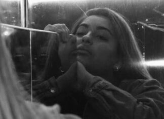 A young woman looks at herself in a scratched mirror, her chin angled up and her expression bored or perhaps sorrowful. The photo is in black and white.