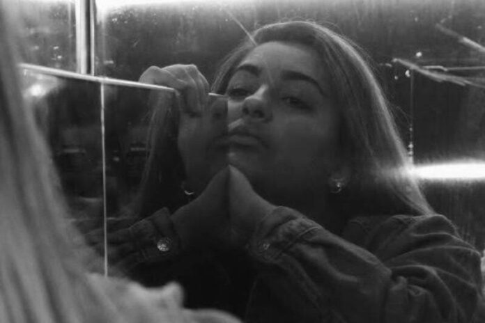 A young woman looks at herself in a scratched mirror, her chin angled up and her expression bored or perhaps sorrowful. The photo is in black and white.
