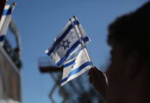A blurred hand holds three small Israeli flags against a blue sky.