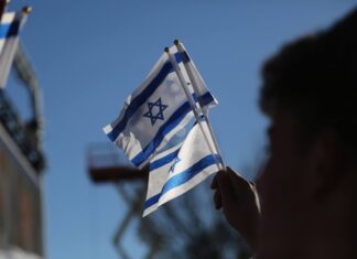A blurred hand holds three small Israeli flags against a blue sky.