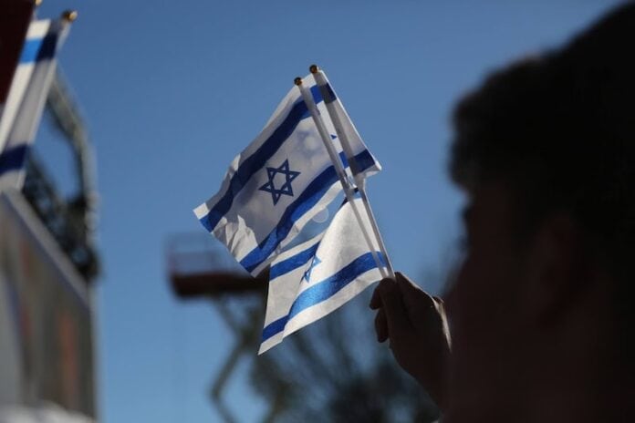 A blurred hand holds three small Israeli flags against a blue sky.