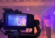 A camera films a group of people in a dimly lit room with purple lighting; the camera display shows two women posing.