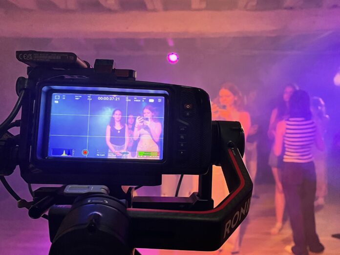 Filmmaker's Perspective A camera films a group of people in a dimly lit room with purple lighting; the camera display shows two women posing.