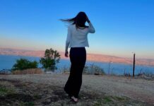 A Simchat Torah to Remember A girl, viewed from behind, looks out over a vibrant blue lake at sunset. Her long brown hair floats in the wind.