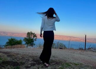 A Simchat Torah to Remember A girl, viewed from behind, looks out over a vibrant blue lake at sunset. Her long brown hair floats in the wind.