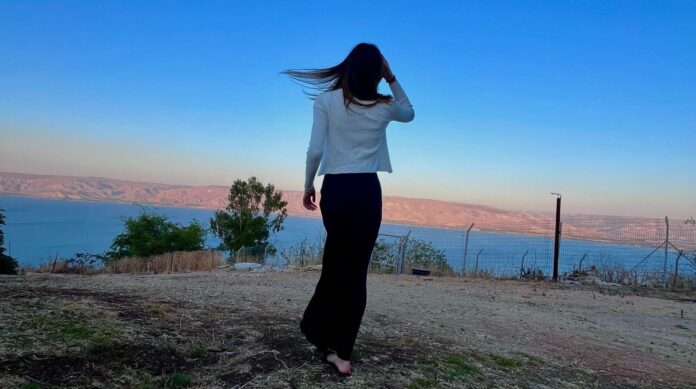 A girl, viewed from behind, looks out over a vibrant blue lake at sunset. Her long brown hair floats in the wind.