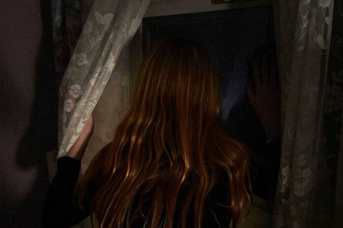 A shiny-haired girl looks out a window with her hand pressed on it.