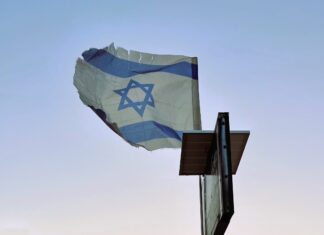 A tattered Israeli flag flies on a post against a pale blue sky.