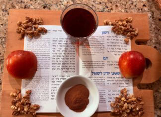 On a cutting board, there is a Haggadah open to "The Four Sons" section. It is surrounded by a cut apple, a cup of wine, a bowl of cinnamon, and walnuts.