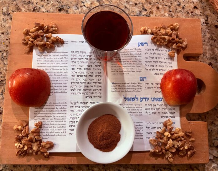 On a cutting board, there is a Haggadah open to "The Four Sons" section. It is surrounded by a cut apple, a cup of wine, a bowl of cinnamon, and walnuts.