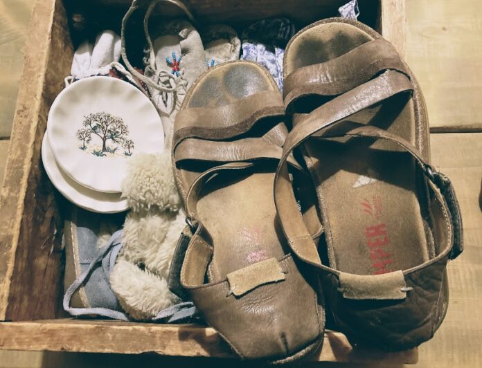 A pair of brown leather sandals, stained with wear, sit atop a wooden crate filled with antique objects.