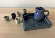 Kiddush Set Naama Raun's Kiddush set includes a blue pitcher with five small individual cups. The pitcher and one of the individual cups sits on a ceramic tray decorated with floral print.
