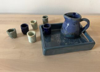 Kiddush Set Naama Raun's Kiddush set includes a blue pitcher with five small individual cups. The pitcher and one of the individual cups sits on a ceramic tray decorated with floral print.