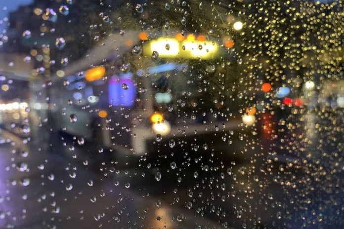 A city bus seen through a pane of glass covered in raindrops. The image is blurry.