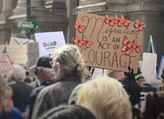 Winter Protesters march near a cardboard sign that says "Migration is an act of courage" surrounded by monarch butterflies.