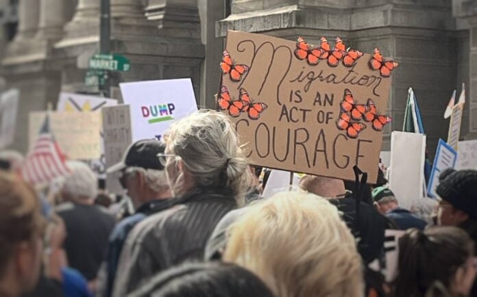 Migration is an Act of Courage Protesters march near a cardboard sign that says "Migration is an act of courage" surrounded by monarch butterflies.
