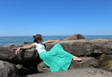 Mermaid A young woman in a turquoise skirt lounges on a rocky outcrop beside a body of water. A lone seagull flies overhead.