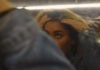 The Way I Look A teen girl is running her fingers through her hair while looking at her reflection in a slightly blurry mirror. Her arm covers one of her eyes and the bottom of her face, leaving only the top of her face visible.